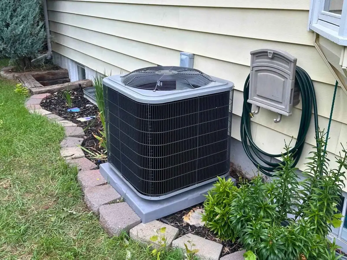 Outdoor central air conditioner on a support pad beside a light-sided home and garden bed.