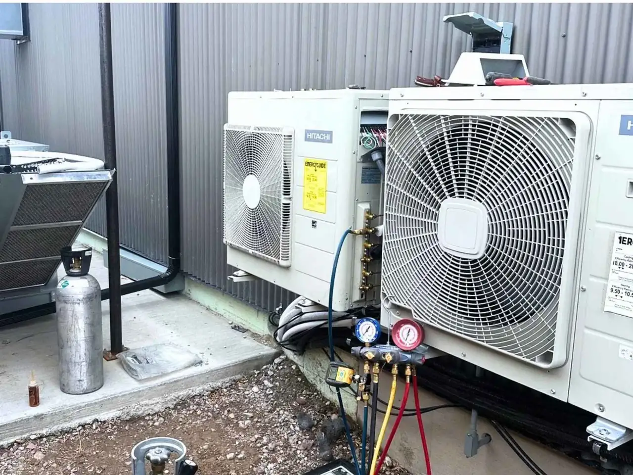 Outdoor HVAC condenser units with connected refrigerant gauges and service hoses beside a metal-sided building.